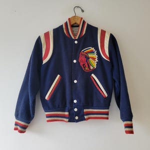Puede incluir: Una chaqueta de béisbol azul marino con rayas rojas y blancas en las mangas y el bajo. La chaqueta tiene una gran imagen bordada de un jefe nativo americano en la parte delantera.