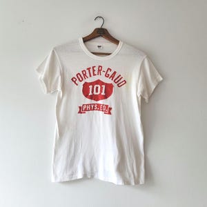 May include: A white t-shirt with a red graphic that reads "PORTER-GAUD 101 PHYS.ED." The shirt is hanging on a wooden hanger. The t-shirt has short sleeves and a crew neck.