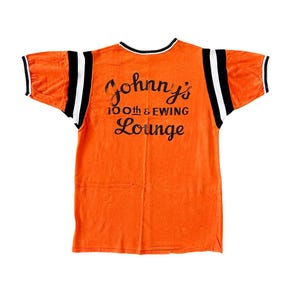 May include: Orange jersey with black and white stripes on the sleeves and shoulders. The back of the jersey features the text "Johnny's 100th & Ewing Lounge" in black lettering. The collar and sleeve cuffs are trimmed in black and white.
