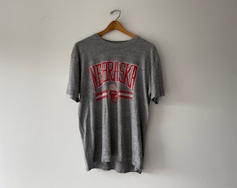 Könnte beinhalten: Graues T-Shirt mit einem roten und weißen "Nebraska"-Motiv und einem Footballhelm.