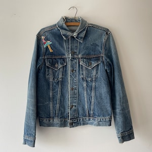 May include: A blue denim jacket with a small embroidered patch on the left chest. The jacket has a classic style with a button-up front and two pockets.