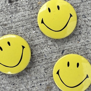 May include: Three yellow smiley face buttons with black outlines. The buttons are on a gray concrete surface.
