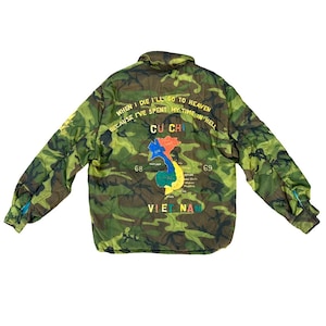 Puede incluir: Chaqueta acolchada de camuflaje con cuello redondo. La parte trasera presenta un mapa colorido de Vietnam con el texto "CU CHI" y la cita "WHEN I DIE I'LL GO TO HEAVEN BECAUSE I'VE SPENT MY TIME IN HELL".