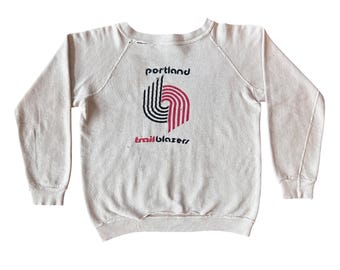 Puede incluir: Sudadera de color crema con el texto "portland" encima de un gráfico de líneas curvas negras y rojas, y la palabra "trailblazers" debajo. La sudadera tiene mangas largas y cuello redondo.