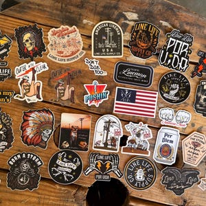 May include: A collection of various vinyl stickers featuring diverse graphic designs, including skulls, utility poles, American flags, and text such as Line Life, Per Diem, and Lineman Club, arranged in a scattered layout on a wooden surface.