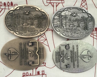 May include: Four belt buckles with a lineman theme. Two are silver and two are brass. The buckles feature a heart with a lineman working on a power line, and the text "Lineman Love Supply Co."
