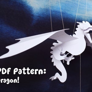 May include: A white paper dragon mobile with wings outstretched. The dragon is hanging from two strings against a blue background. The text "PDF Pattern: Dragon!" is at the bottom of the image.