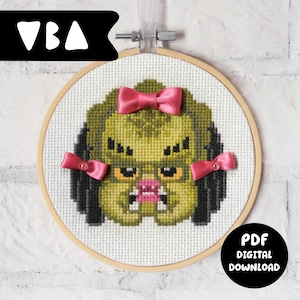 May include: A cross-stitch pattern in a circular wooden hoop featuring a pixelated green creature with black dreadlocks, yellow eyes, and white fangs. The creature is decorated with three small pink satin bows attached to its head and sides.