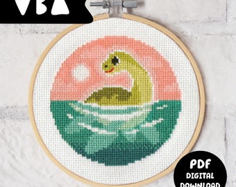 May include: A cross-stitch pattern in a circular wooden embroidery hoop featuring a green dinosaur-like creature emerging from dark teal water against a light pink sky with a white circular sun. The text PDF DIGITAL DOWNLOAD is visible.