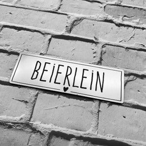 Könnte beinhalten: Ein silbernes Metallschild mit dem Wort "BEIERLEIN" in schwarzer Schrift. Unter dem Wort befindet sich ein kleines Herzsymbol.