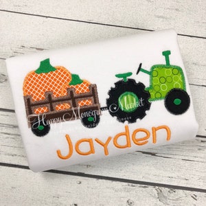 May include: A white shirt featuring an embroidered design of a green tractor pulling a brown wooden wagon filled with orange pumpkins. The name Jayden is embroidered in orange below the tractor and wagon. The design has a visible stitched edge.
