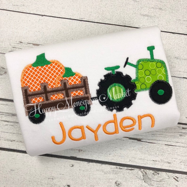 May include: A white shirt featuring an embroidered design of a green tractor pulling a brown wooden wagon filled with orange pumpkins. The name Jayden is embroidered in orange below the tractor and wagon. The design has a visible stitched edge.