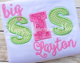 May include: A white t-shirt featuring embroidered applique text that reads big SIS in a combination of pink script, a pink patterned block letter I, and green grid-patterned S letters, with the name Layton embroidered in pink script below.