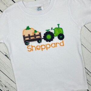 May include: A white short-sleeved t-shirt featuring an embroidered design of a green tractor pulling a brown wagon filled with orange pumpkins. The name Sheppard is embroidered in orange below the tractor. The shirt has an ARB Blanks neck label.