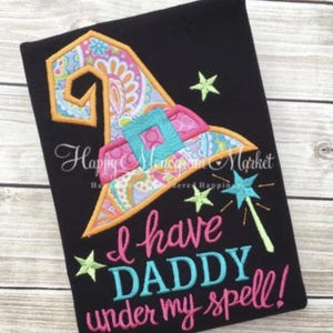May include: A black t-shirt featuring an embroidered witch hat with a colorful paisley and floral pattern, a bright blue buckle, and a star-tipped wand. The text reads, "I have DADDY under my spell!" in pink and blue embroidered lettering.