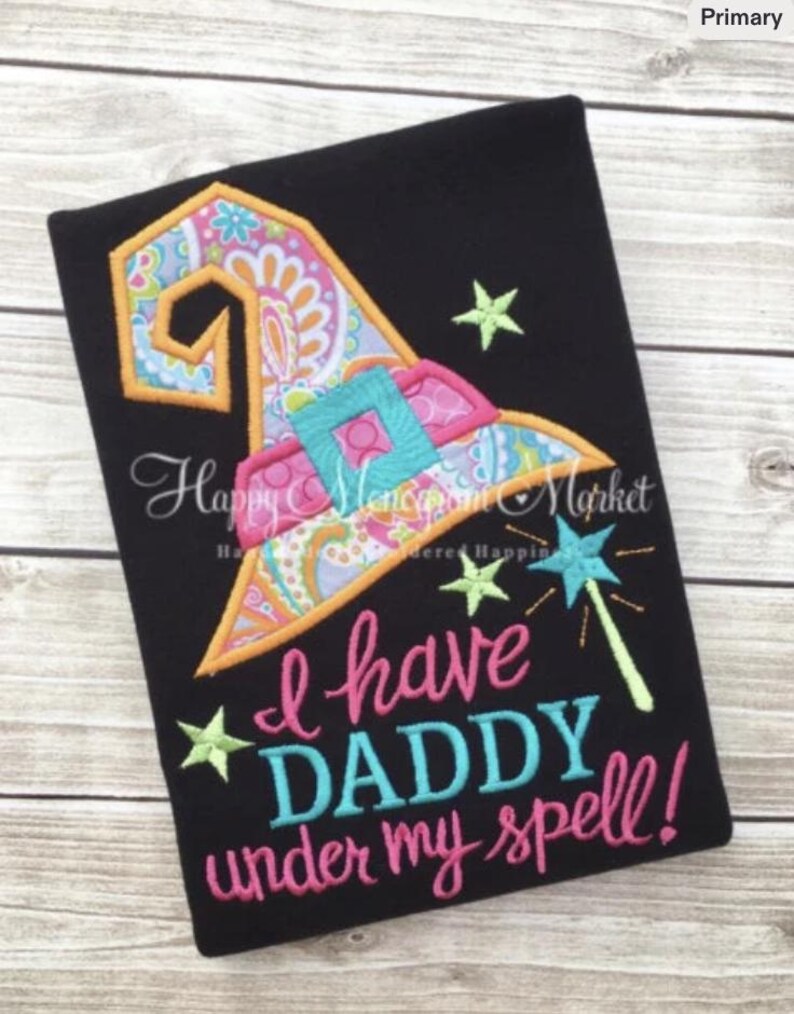 May include: A black t-shirt featuring an embroidered witch hat with a colorful paisley and floral pattern, a bright blue buckle, and a star-tipped wand. The text reads, "I have DADDY under my spell!" in pink and blue embroidered lettering.