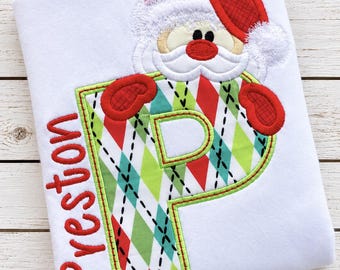 May include: A white t-shirt featuring an embroidered letter P with a red, green, and white argyle pattern. A Santa Claus figure with a red hat and white beard is positioned above the letter. The name Preston is embroidered in red on the left side.