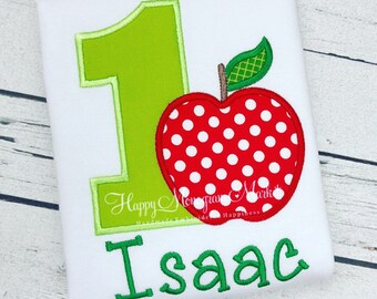 May include: A white shirt featuring an embroidered lime green number one, a red apple with white polka dots, and the name Isaac in green letters. The design includes a green leaf on the apple stem and the text Happy Monogram Mark.