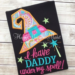 May include: A black shirt featuring an embroidered witch hat with a colorful paisley and floral pattern, a teal buckle, and three lime green stars. The text reads "I have DADDY under my spell!" in pink and teal, accompanied by a teal magic wand.