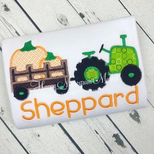 May include: A white shirt featuring an embroidered green tractor pulling a brown wagon filled with orange pumpkins. The name Sheppard is embroidered in orange letters below the tractor and wagon design, which includes a decorative stitch pattern.