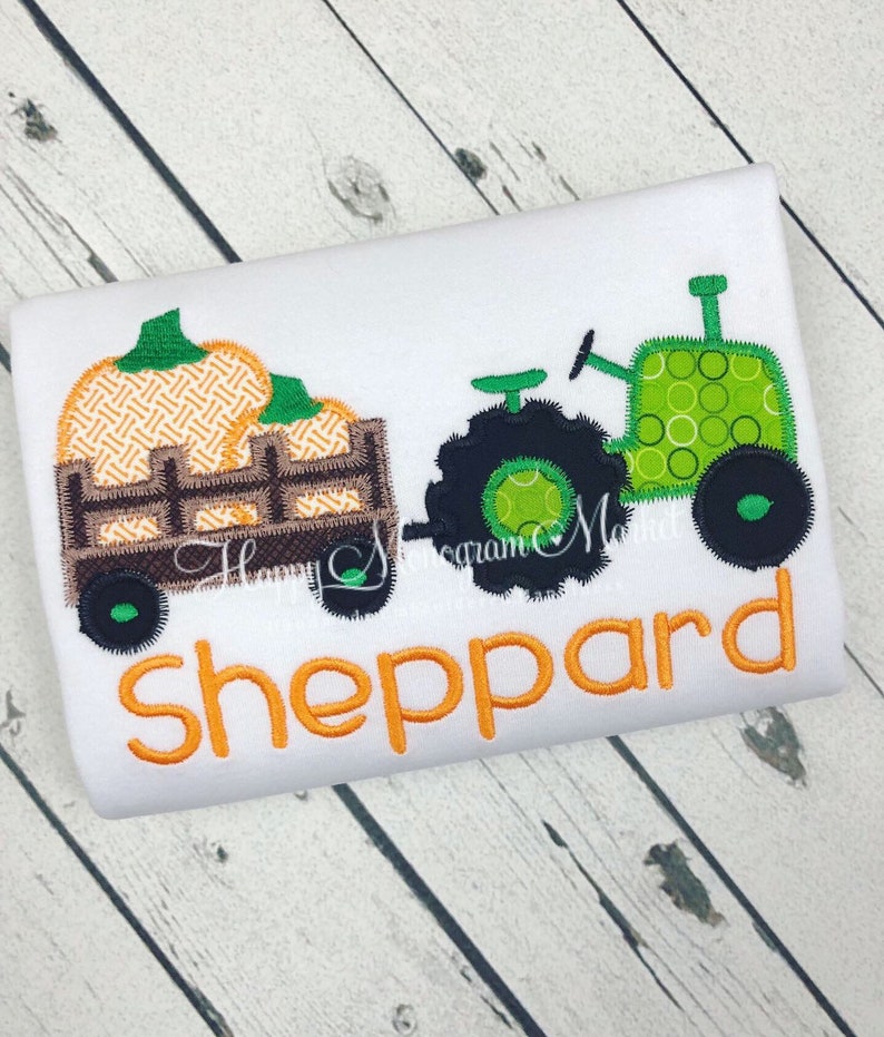 May include: A white shirt featuring an embroidered green tractor pulling a brown wagon filled with orange pumpkins. The name Sheppard is embroidered in orange letters below the tractor and wagon design, which includes a decorative stitch pattern.