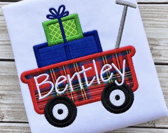 May include: A white fabric square featuring an embroidered red plaid wagon with black wheels and a grey handle. The wagon carries a blue rectangular gift and a green gift with a yellow grid pattern and blue bow. The name Bentley is stitched in white.