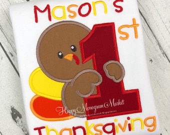 May include: A white shirt featuring an embroidered design of a brown turkey holding a large red number one. The text reads Mason's 1st Thanksgiving in red and orange thread. The design includes yellow and orange tail feathers and a small watermark.