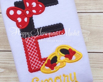 May include: A white shirt featuring a large embroidered letter E in black and red plaid, accented with a red and white polka dot bow. Two yellow shoes with red bows are stitched near the base, and the name Emory is embroidered in yellow below.