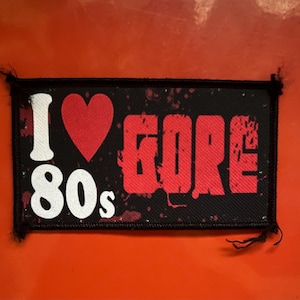May include: A rectangular fabric patch with a black background featuring the white text "I" and "80s" alongside a red heart symbol and the red text "GORE". The design includes red splatter patterns and is finished with a black stitched border.