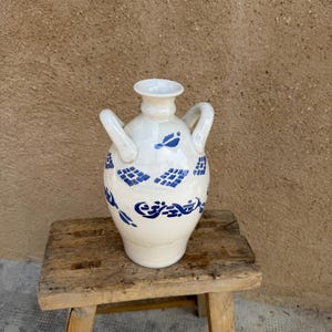 Könnte beinhalten: Eine weiße Keramikvase mit zwei Henkeln und einem ausgestellten Rand. Die Vase ist mit blauen geometrischen und floralen Mustern verziert. Sie steht auf einem kleinen, verwitterten Holzschemel. Der Hintergrund ist eine strukturierte Wand in neutralen Tönen.