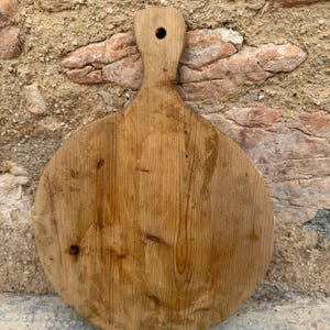 Puede incluir: Una tabla de cortar redonda de madera con un asa y un agujero para colgar. La tabla es de color marrón claro con vetas de madera visibles y algunos nudos oscuros. El asa tiene forma redondeada. El fondo es una pared texturizada de color beige.