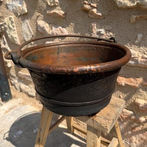 Peut inclure: Un chaudron en cuivre vieilli, avec une patine sombre et ancienne, muni d'une anse robuste. Le pot est posé sur un petit tabouret en bois rustique. L'intérieur du chaudron montre des signes d'utilisation.