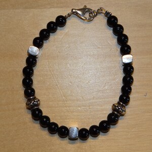 Op de afbeelding: Een zwarte kralen armband met zilveren accenten. De armband heeft een sluiting en is gemaakt van ronde kralen met een gladde afwerking.