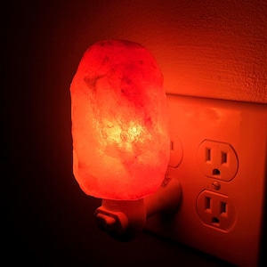May include: A pink Himalayan salt lamp plugged into a standard electrical outlet. The lamp is glowing a warm orange color.