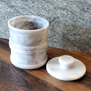 May include: A white marble container with a lid. The container has a gray marbled pattern and a slightly raised rim. The lid has a small knob on top.