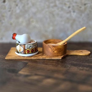 May include: A wooden serving board with two small ceramic bowls. One bowl is white with a red, yellow, and green design and the text "Epices". The other bowl is brown and has a wooden spoon inside.