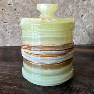 May include: A cylindrical, lidded jar made of layered, translucent green onyx. The jar features bands of brown, orange, and white, with a rounded base and a small, knob-like handle on the lid. The jar is approximately 4 inches tall.