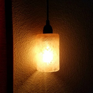 May include: A warm, glowing salt lamp with a textured, orange-brown finish hangs from a black cord against a textured, off-white wall.