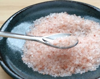 May include: A close-up of a small, shallow, dark blue ceramic dish filled with pink Himalayan salt. A silver spoon is scooping up the salt.
