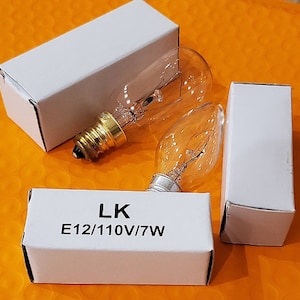 May include: Two clear glass light bulbs with a candelabra base, next to two white rectangular boxes. The text "LK E12/110V/7W" is printed on one of the boxes. The bulbs have a flame-like shape and are on an orange background.