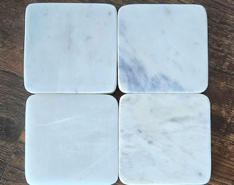 May include: Four white marble coasters with a light gray veining pattern. The coasters are square shaped and have a smooth, polished surface.