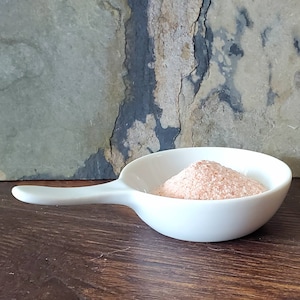 May include: A small white ceramic bowl with a handle, filled with a mound of pink Himalayan salt.