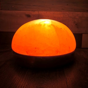 Może przedstawiać: Okrągła, pomarańczowa lampa solna z Himalaj&oacute;w z czarną podstawą. Lampa jest włączona i świeci.