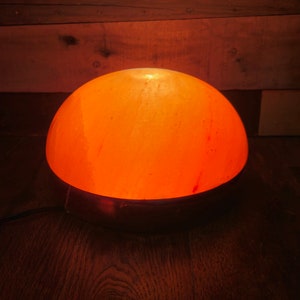 Może przedstawiać: Okrągła, pomarańczowa lampa solna z Himalaj&oacute;w z metalową podstawą. Lampa jest włączona i świeci.