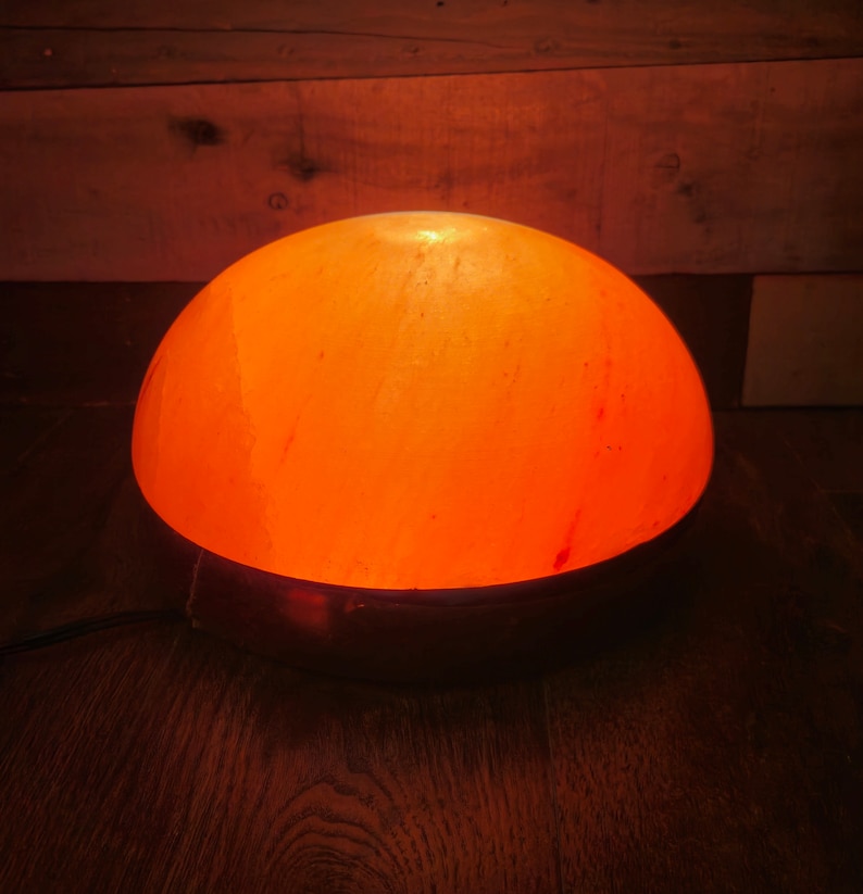 Może przedstawiać: Okrągła, pomarańczowa lampa solna z Himalaj&oacute;w z metalową podstawą. Lampa jest włączona i świeci.