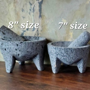 Może przedstawiać: Dwa moździerze i tłuczki molcajete z szarego wulkanicznego kamienia, jeden o średnicy 20 cm, a drugi 18 cm. Moździerze znajdują się na drewnianej powierzchni.