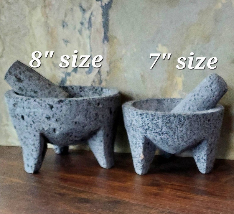 Może przedstawiać: Dwa moździerze i tłuczki molcajete z szarego wulkanicznego kamienia, jeden o średnicy 20 cm, a drugi 18 cm. Moździerze znajdują się na drewnianej powierzchni.