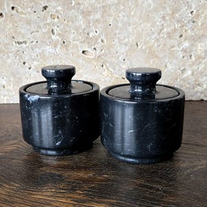 May include: Two matching black marble jars with lids. The jars are cylindrical with a rounded base and a small, round knob on top of the lid. The marble has a natural, veined pattern. The jars are displayed on a dark wooden surface.