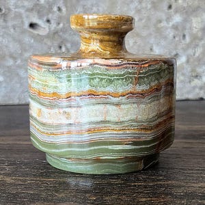 May include: A decorative onyx vase with a cylindrical body and a flared neck. The vase features bands of green, brown, orange, and cream colors. The base is a solid green color. The vase is approximately 4 inches tall.