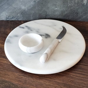 May include: White marble cheese board with a small white marble bowl and a silver cheese knife.